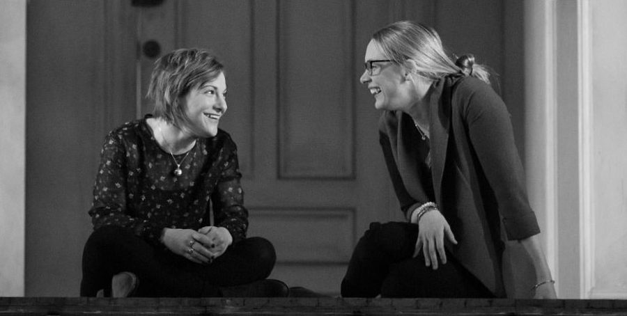 Black and white photo of two women smiling at each other as they sit cross-legged in front of a door