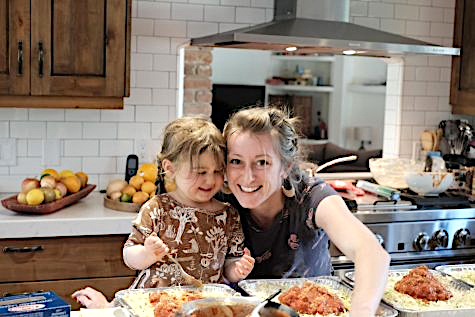 Lasagna Love founder Rhiannon Menn (right) leans in near her young daughter as they both spoon sauce into lasagna pans. The organization was founded through Menn's desire to help families who were in need during the pandemic and has now expanded to anyone in a hardship. 